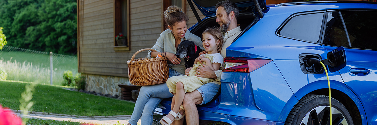 Man, Woman, and younger girl with an elder black dog sitting in the open trunk of an electric vehicle while charging.