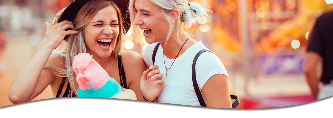 Two young women laughing and sharing cotton candy at a fair.